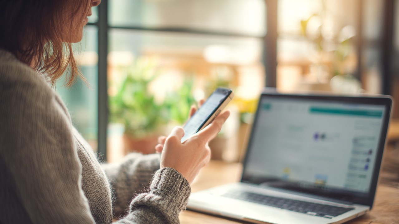 Close-up of a woman using a smartphone and laptop simultaneously to browse an online education platform in a cozy home setting with natural daylight.