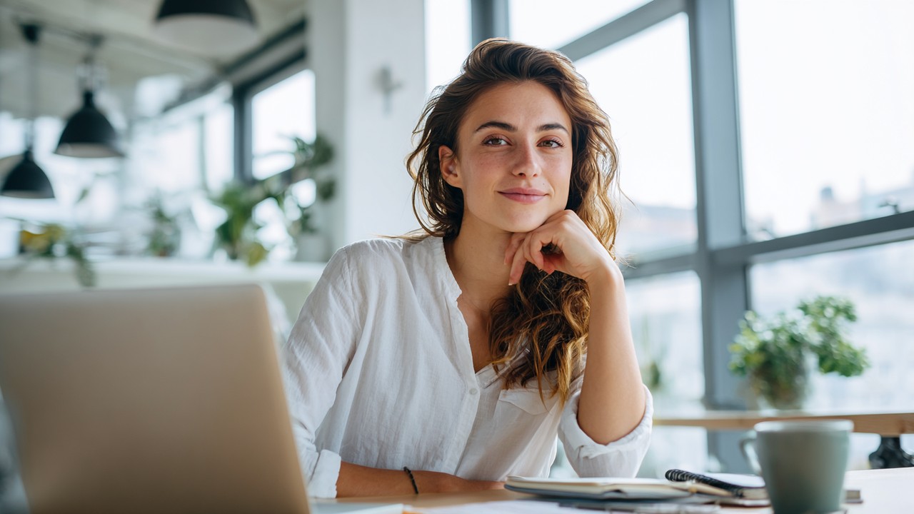 Confident young woman working on laptop in bright modern home office with notebooks, coffee, and natural light.