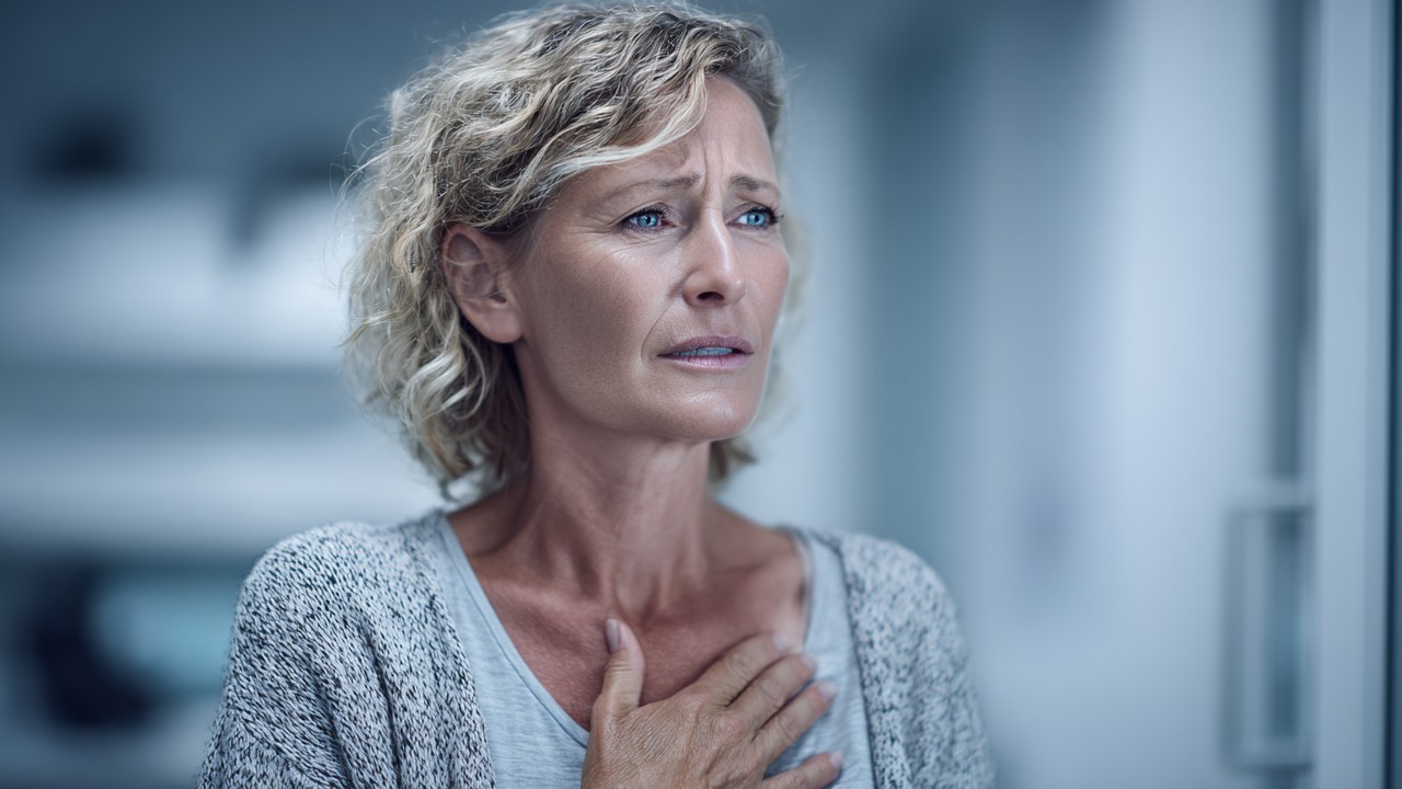 Middle-aged Turkish woman looking concerned and holding her chest in a clinical setting, highlighting heart health and stress effects.