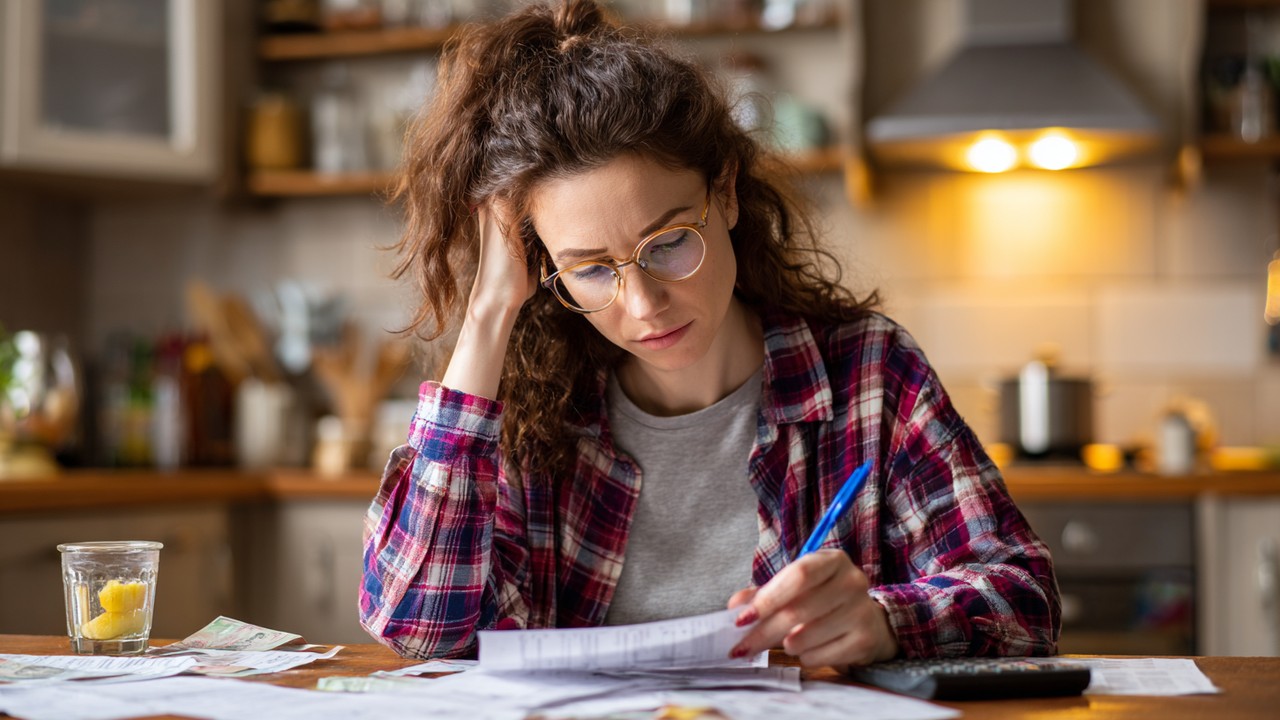 Turkish woman reviewing monthly expenses with pen and notebook at kitchen table, surrounded by bills and receipts in a warm home setting.