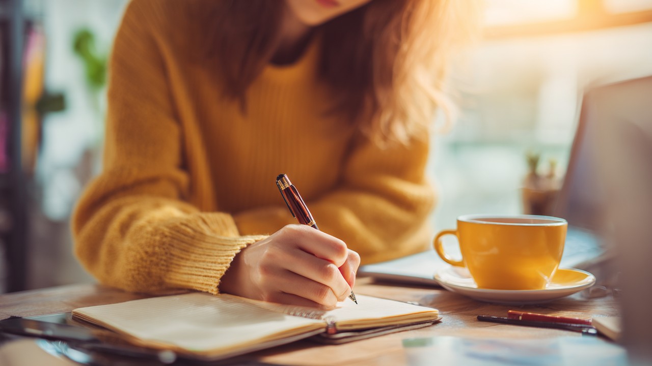Turkish woman writing financial goals on a notebook at a bright desk with a laptop and tea, symbolizing financial planning and future security.