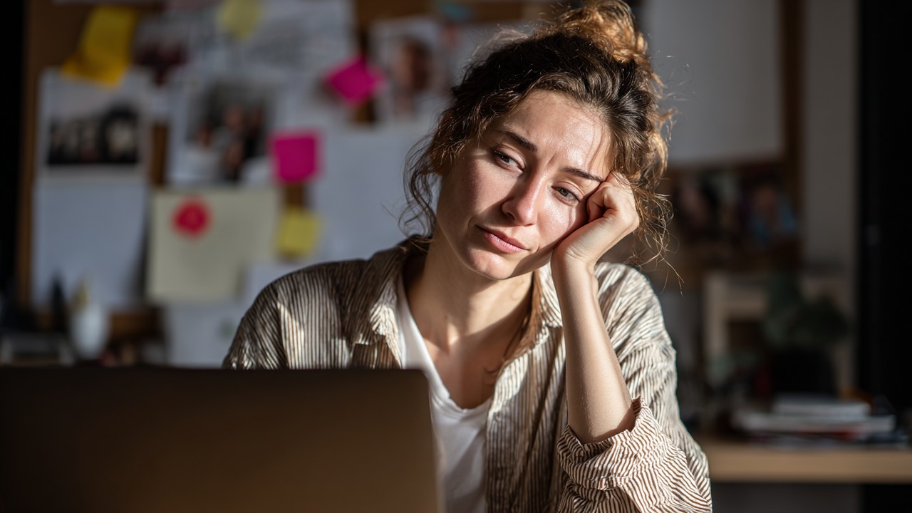 Turkish woman studying online at her desk with family photos, notes, and laptop, showing focus and concern in natural lighting.
